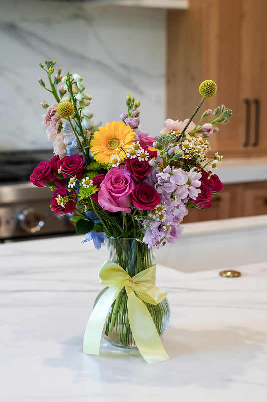 Bouquet of colorful flowers in a clear vase with a yellow ribbon on a kitchen counter.